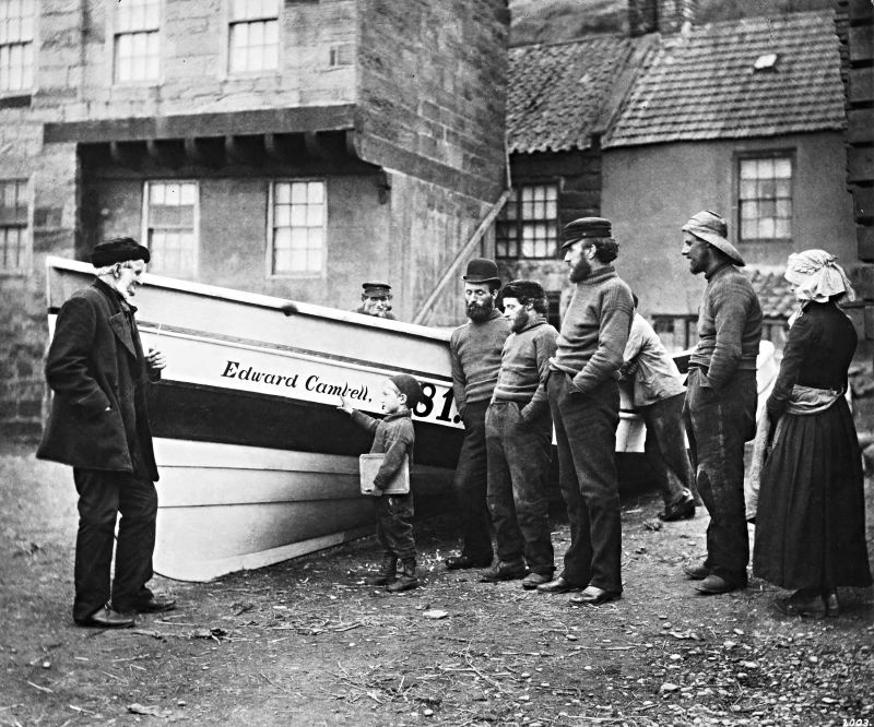 Frank Meadow Sutcliffe (1853-1941) documented in a masterly manner daily life in Whitby along the North Sea coast and the surrounding district – he had begun his career as a studio photographer, which remained his most significant source of work. Local inhabitants were photographed in his studio, and many visitors wanted their pictures taken during their holidays by the sea. But it became his close observations of the Whitby fishing community that made him famous. One such photograph of interest is this picture from Whitby of the boat ‘Edward Cambell’ and a group of local fishermen with family members in the 1890s. In the context of this particular essay, notice the woman’s traditional bonnet, with its raised longitudinal bands, which enabled the wearer to carry baskets on her head. At the same time, the extension at the back prevented water from running down her neck. Whilst most of the men and the small boy wore woollen knitted ganseys. (Courtesy: Private Collection. Public Domain). Photo: Frank Meadow Sutcliffe.