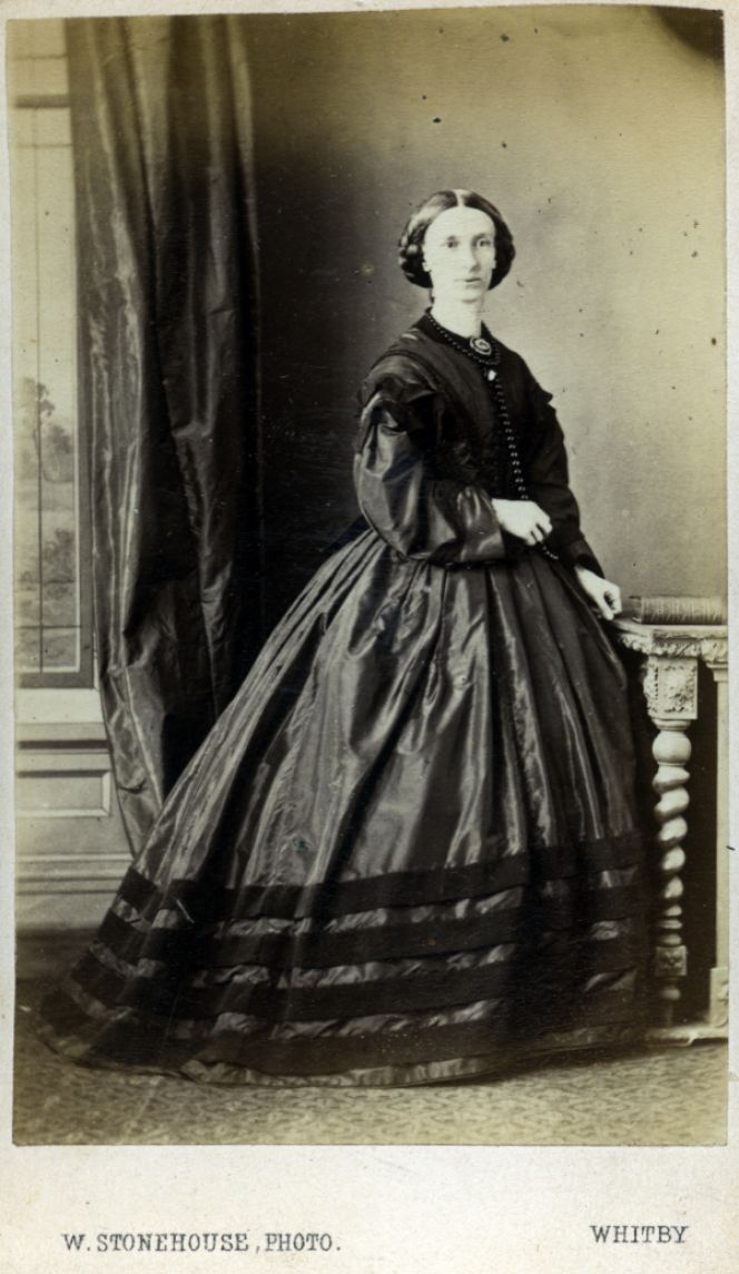 This lady is dressed in a dark coloured satin dress decorated with broad velvet ribbons. A sort of dress which  requires a professional dressmaker’s hand to reach perfection in cut and sewing. The photograph is taken in  1866 by the photographer W. Stonehouse. The Pier Portrait Rooms in Whitby. Courtesy of:  Whitby Literary & Philosophical Society, Whitby Museum, Photographic Collection.
