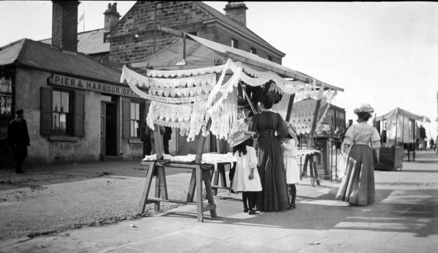 In this early 20th century photograph a display of different breadths of lace has attracted the attention of a well-dressed lady with two small daughters at her side. It seems that this lace stall operated on this spot during the summer months up to the outbreak of the First World War. (Courtesy of: Whitby Museum/Whitby Lit. & Phil. Photographic Collection, W 4394.)