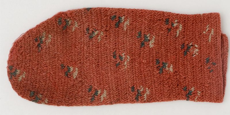 This woollen mitten, dated to circa 1820s-1850s, is unusual in style with its regularly placed motifs in white and green in a madder red ground. That is to say, the scattered decoration was part of the nålbindning technique itself. In contrast to most decorative needle-bound mittens from 19th century Sweden, where designs instead used to be added afterwards with colourful embroidery – like illustrated below. (Courtesy: The Nordic Museum, Stockholm, Sweden. NM.0221366. | One single extant mitten. Sunnerbo district, Småland province, Sweden. Digitalt Museum).