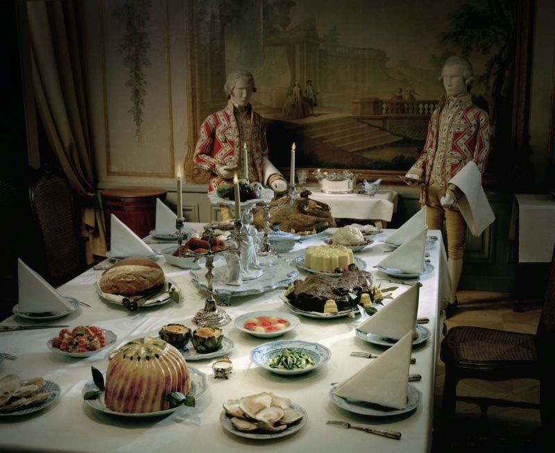 In stark contrast to the everyday life of the illustrated linen washerwoman, this reconstruction of an elegantly laid table in a Swedish home of the nobility in mid-18th century, demonstrates how the linen napkins and large-size tablecloth were important parts of the festive meal. One can reflect on that the extensive width as well as length of the fabric was necessary for the tablecloth to reach the floor at all four sides of the table. However, one may notice that the choice of linen tablecloth for this reconstruction has been woven in full width, which was not customary around 1750. Even if the hand-woven cloth in damask or diaper had been purchased from a well-established linen manufacturer, the loom widths were not wide enough, so two exact pieces of fabric had to be finished off with a long seam to obtain the desired width – traditionally in the finest stitching to be as invisible as possible. (Courtesy of the Nordic Museum, Stockholm, Sweden. NMA.0000533, The Exhibition “Dukade bord”. Digitalt Museum).