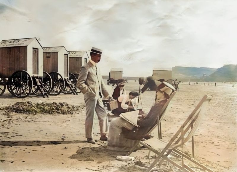 Man in a light-coloured tailored suit and straw boater hat, appropriate beach dress for c.1905-1910. In company with a lady comfortably seated in a deckchair with her newspaper. The couple had chosen to carry their chairs quite a bit from the cliffside at this ebb phase; maybe they were planning to change into bathing suits in one of the bathing machines when the water flowed in. Overall, the popularity of this stretch of beach is not only its beauty and vicinity of the town’s facilities but also that even if the difference between the ebb and flow of the tide can be up to five and a half meters in Whitby, there is always at least a small narrow piece of dry land even at the highest tide. (Courtesy: Whitby Museum, Photographic Collection, C466 & added modern digital colouring. Unknown photographer).