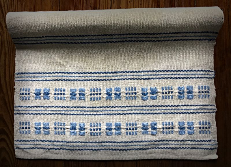 Some of my own weaving experience with this technique dates back many years, in the form of six table mats (26 cm x 40 cm) in the “munkabälte” variation on four shafts and four treadles. For the warp, unbleached cotton yarn 20/2, and for the weft, bleached linen yarn 16/1 and two blue shades, were used. Two treadles were used for the shuttle main weft and two for the shuttle brocading weft. The table mats have got some wear and tear over the years, but are still useful after four decades.| Material and weaving notes: Malmö Handicraft Organisation in the 1980s. (Private ownership). Photo and woven table mat: Viveka Hansen, The IK Foundation.